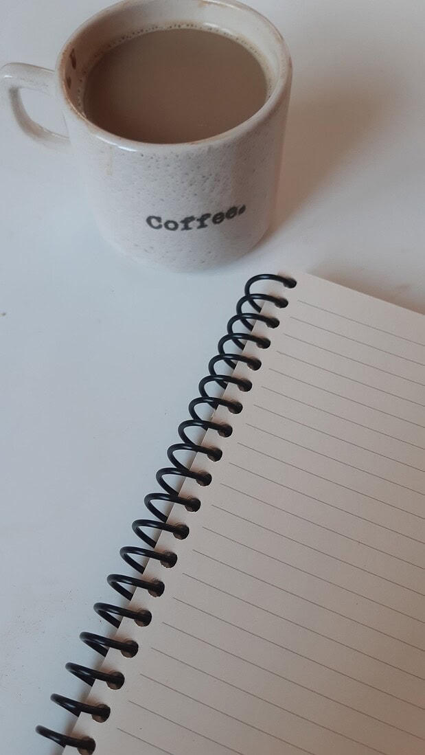 A cozy setup with a coffee mug and spiral notebook on a clean white table, perfect for writing or journaling.