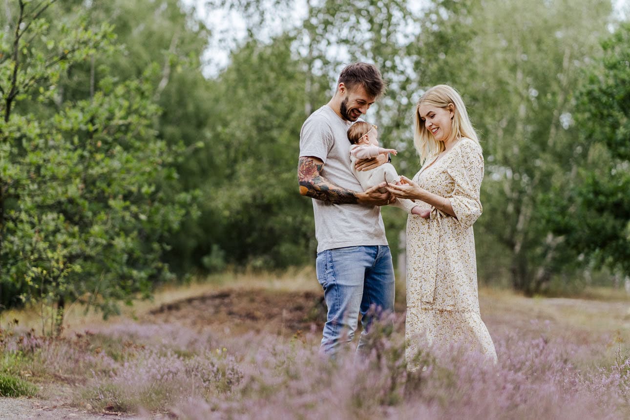 Glückliche Familie mit Baby in der Heide in Münster, umgeben von blühenden Pflanzen und strahlendem Sonnenschein.