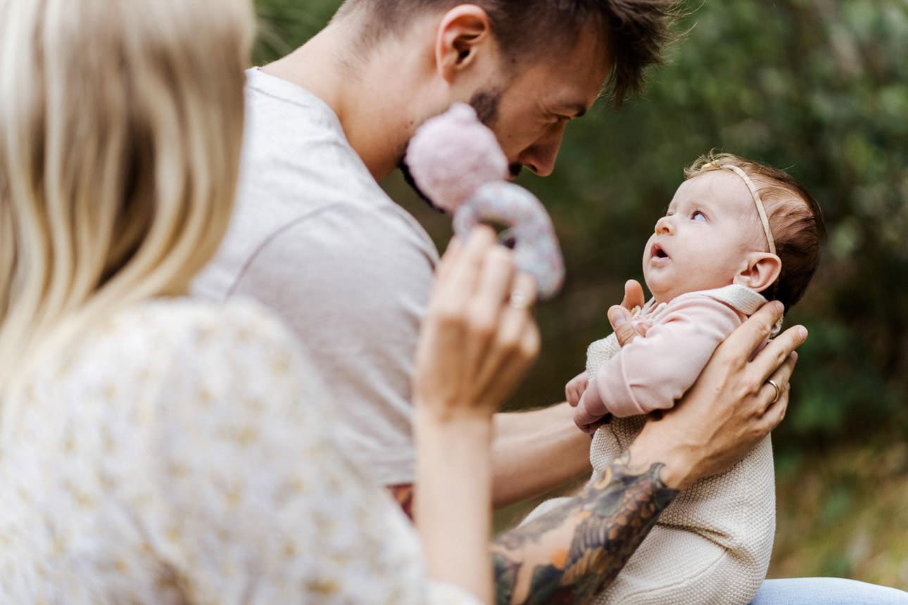 Ein Vater hält seine Tocher auf dem Arm. Die Mutter winkt mit einem Kuscheltier.