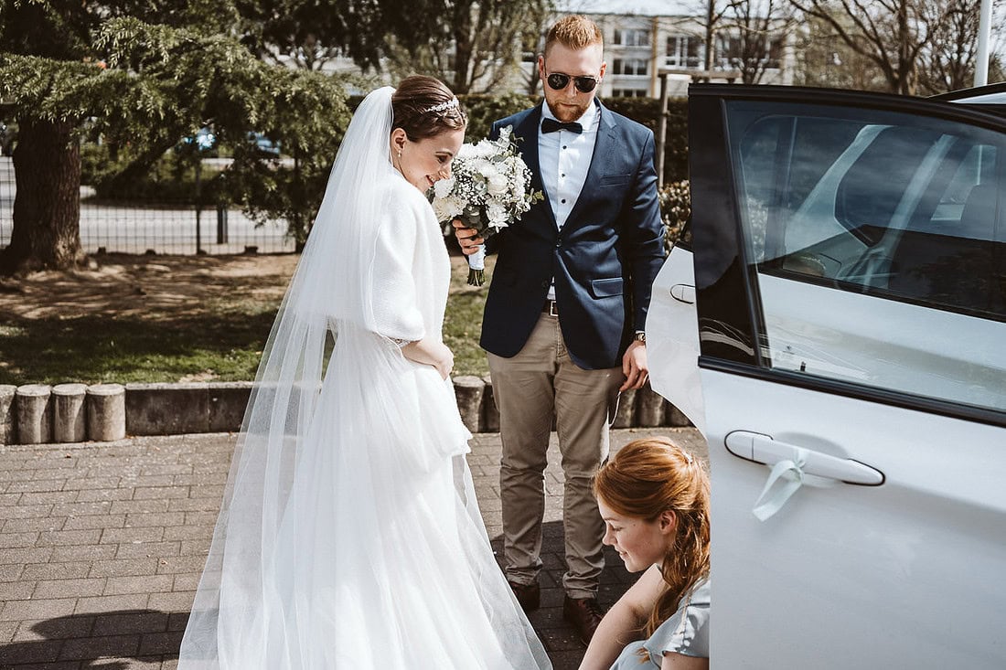 Braut steigt aus dem Auto, während die Trauzeugin der Braut mit dem Kleid hilft.