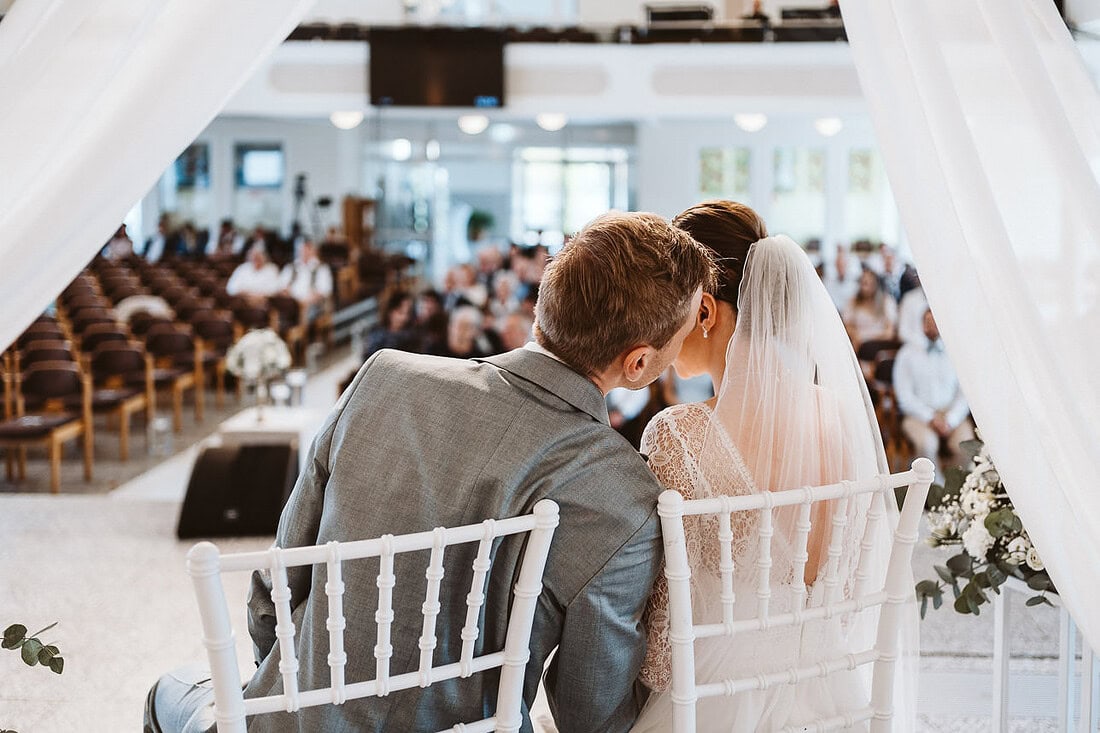 Ein Hochzeitspaar sitzt unter einem Traurigen. Der Bräutigam flüstert der Braut etwas ins Ohr.