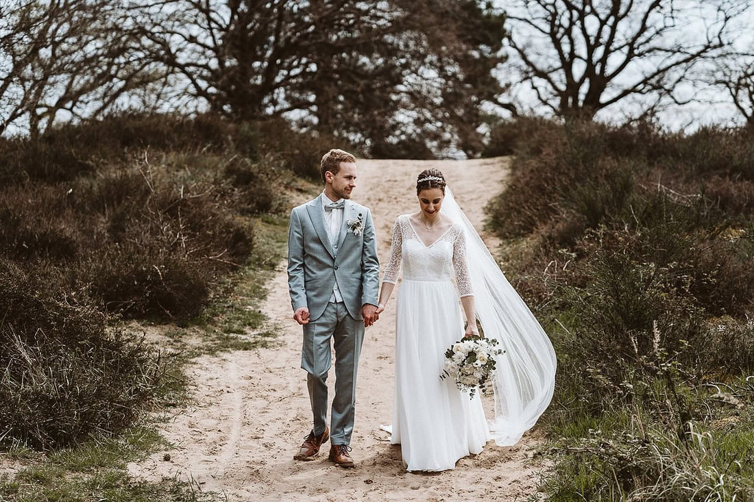 Hochzeitspaar spaziert Hand in Hand einen Pfad in der Heide entlang.