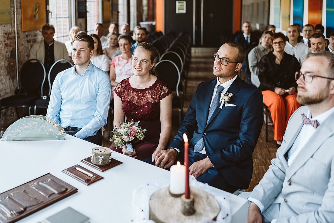 Ein Hochzeitspaar sitzt mit den Trauzeugen im Standesamt. Im Hintergrund sind die Gäste zu erkennen.