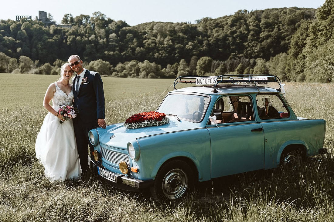 Ein Hochzeitspaar umarmt sich und steht neben einem Trabant mit Blick auf die Burgruine in Windeck.