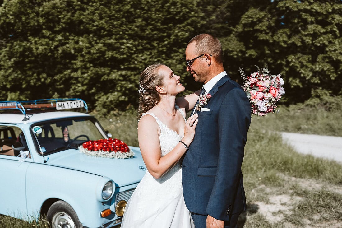 Eine Braut umarmt ihren Bräutigam und hat ihren Brautstrauss in der Hand. Im Hintergrund steht ein blauer Trabant.