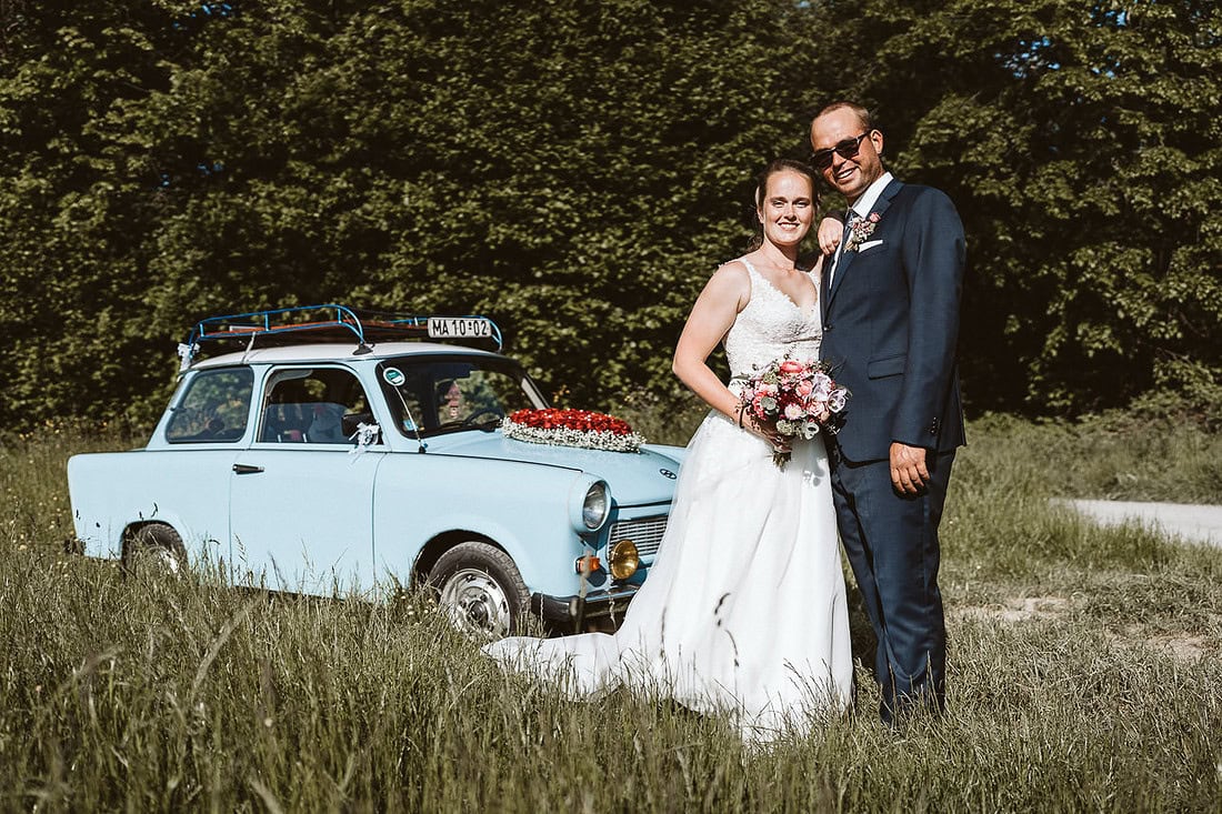 Ein Hochzeitspaar steht auf einer Wiese und blickt in die Kamera. Im Hintergrund steht ein blauer Trabant.