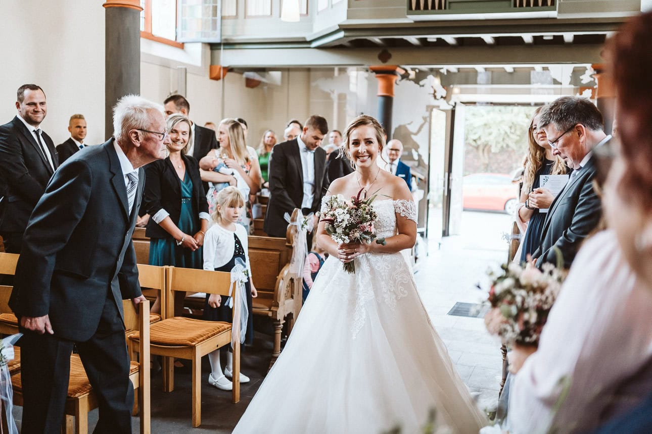 Eine Braut schreitet durch den Mittelgang der Kirche nach vorne zum Altar. Alle Gäste schauen sie an und sind fröhlich.