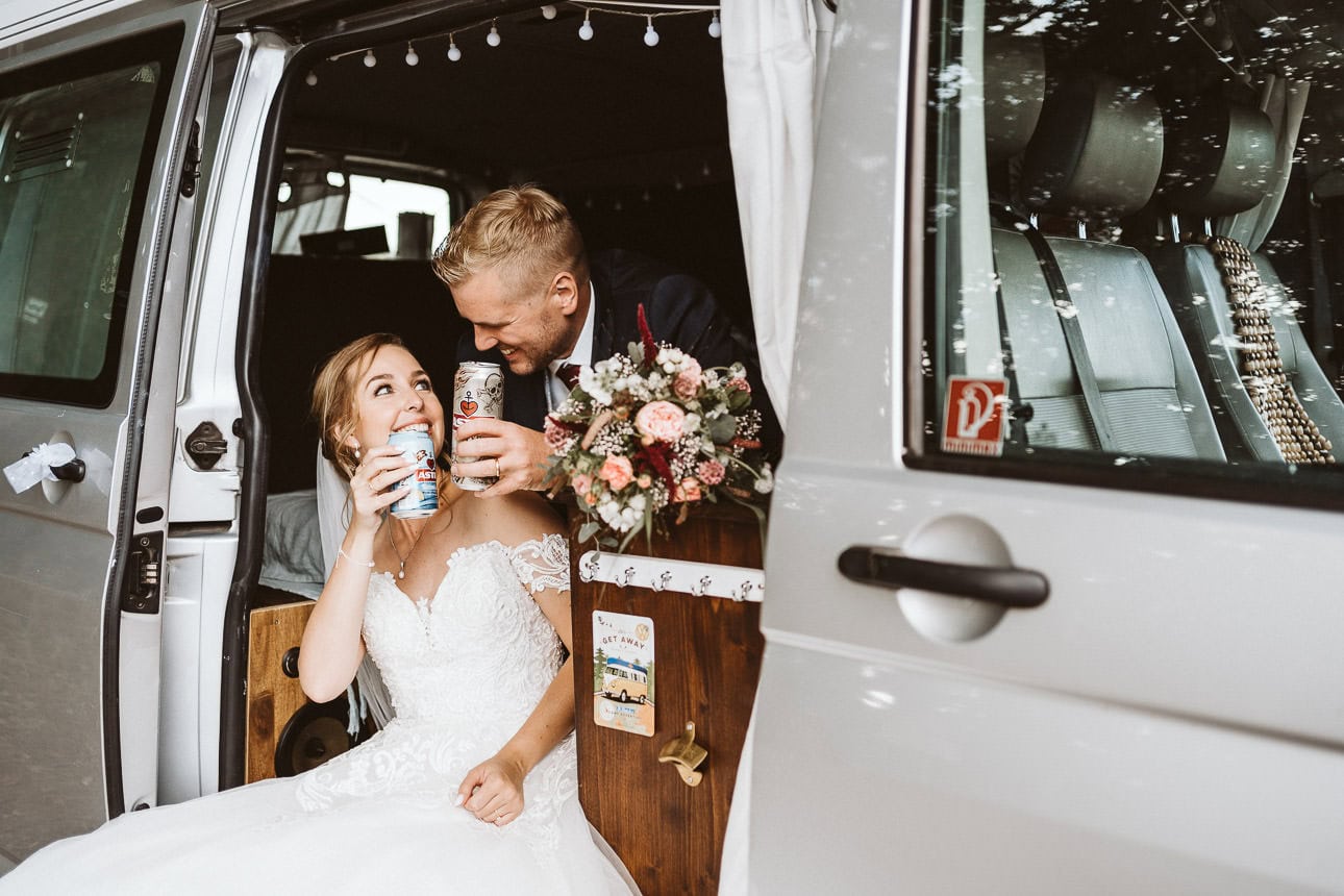 Ein Hochzeitspaar sitzt in einem selbst ausgebauten Camper Van und trinkt ein Dosenbier.