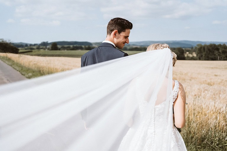 Ein Hochzeitspaar steht an einem Feldrand und schaut sich an. Der Schleier der Braut weht im Wind.