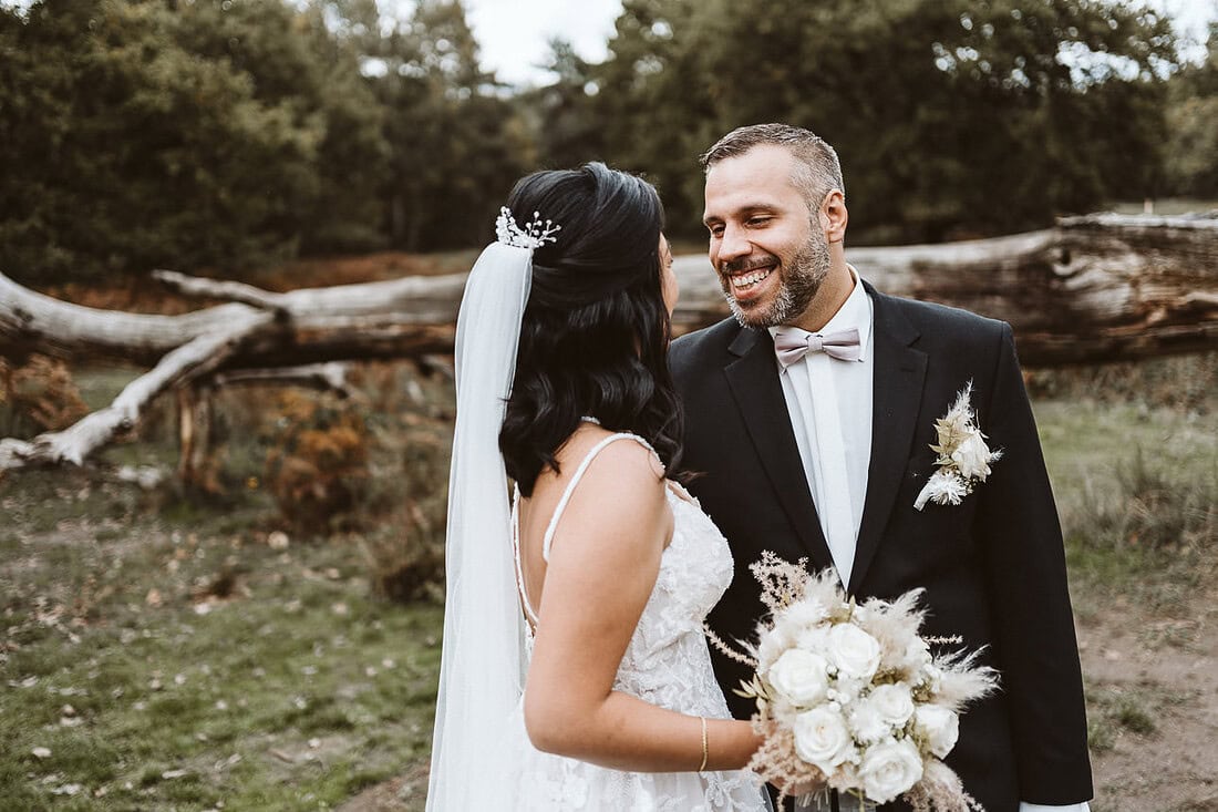 Ein Hochzeitspaar steht vor einem grossen Baumstamm und lächelt sich an.