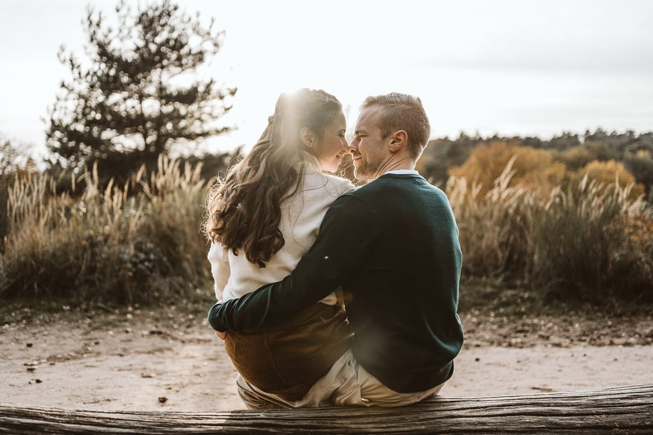 Ein Paar sitzt im Gegenlicht auf einem Baumstamm, schaut sich an und lächelt dabei.