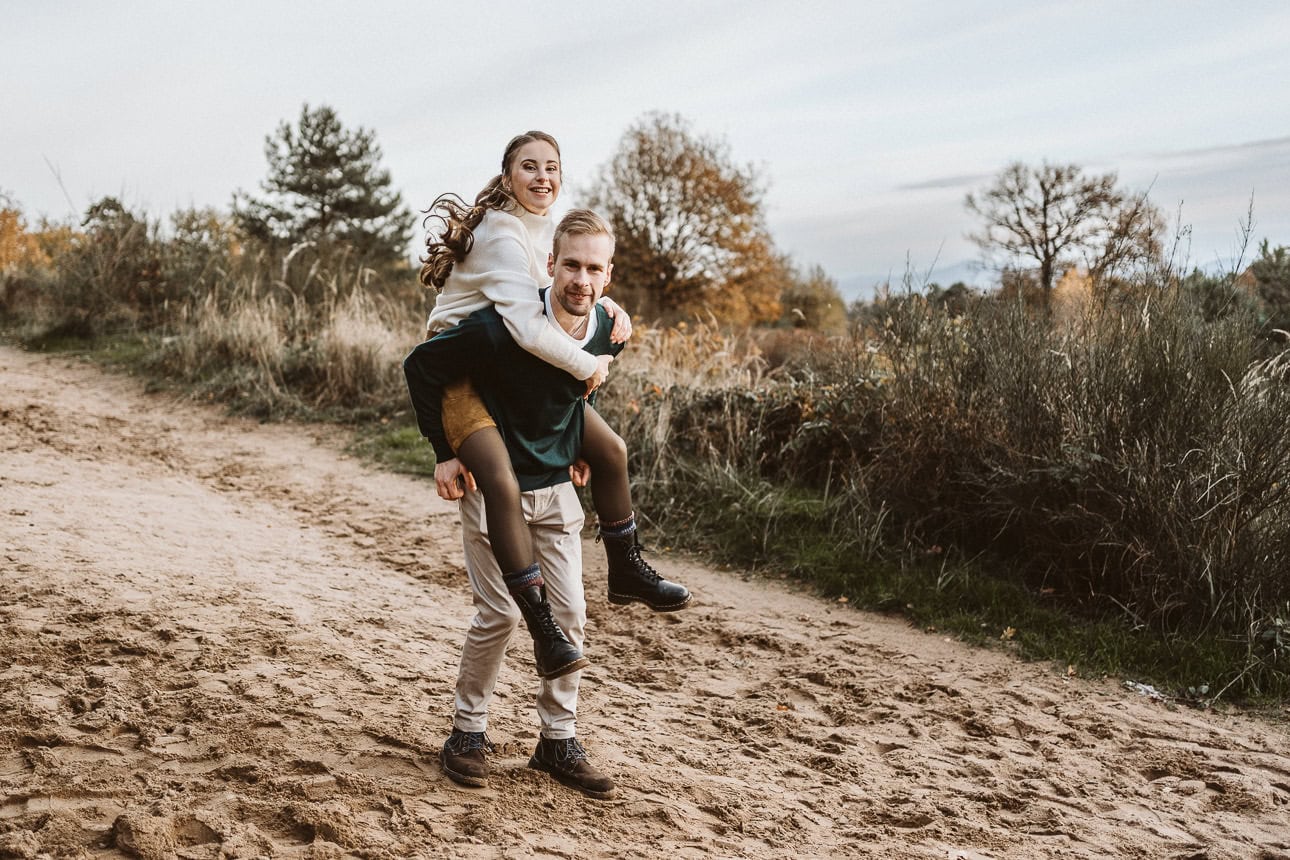 Ein Paar läuft auf einem Weg durch die Heide. Er trägt seine Verlobte auf dem Rücken.